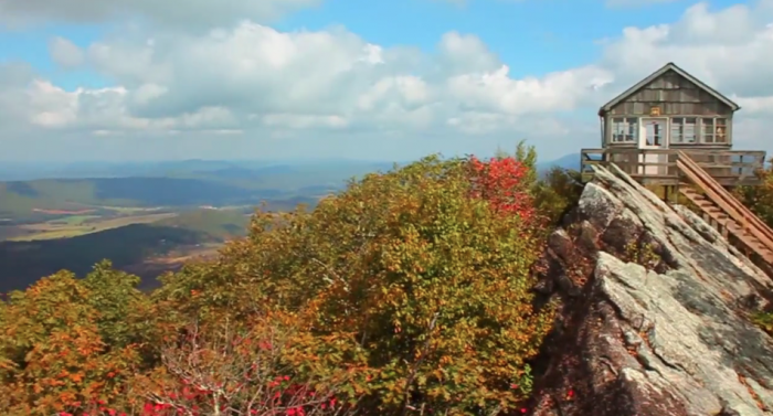 A rustic fire lookout tower perched on a rocky mountain summit surrounded by colorful autumn foliage and sweeping valley views.
