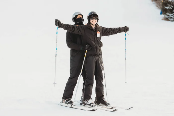 A woman on skis, holing poles, with her arms open wide while a man, also on skis, holds her from behind. -- Winterplace Ski Resort
