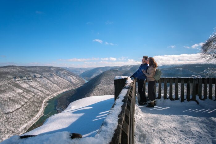 A couple stands on a snow covered wooden deck overlook the mountains and a river in the valley