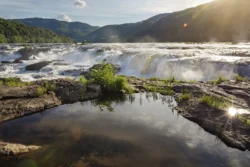 Wide waterfall cascades over a rocky ledge with mountains and trees in the background under bright sunlight