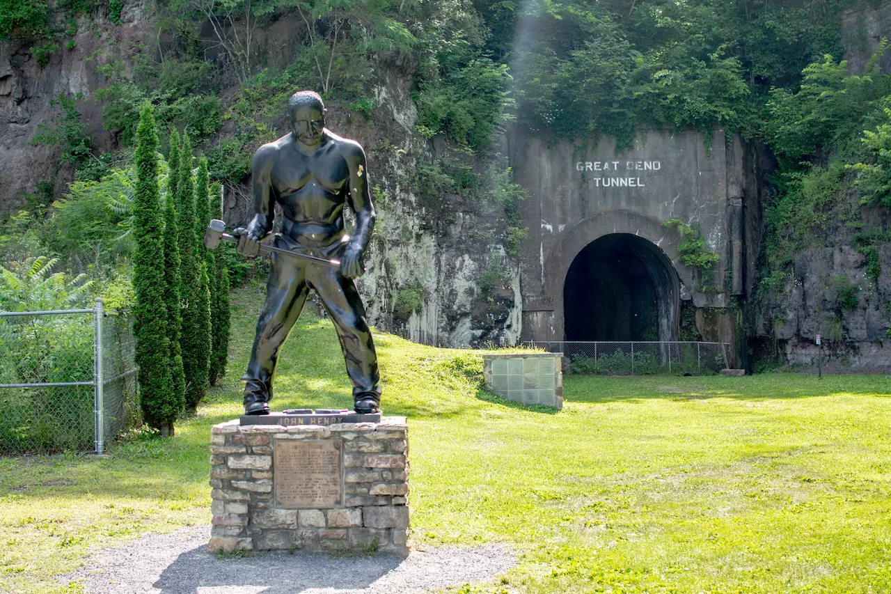 Bronze statue of John Henry with a hammer on a stone pedestal near the forested entrance of the Great Bend Tunnel, with sunlight filtering through the trees.