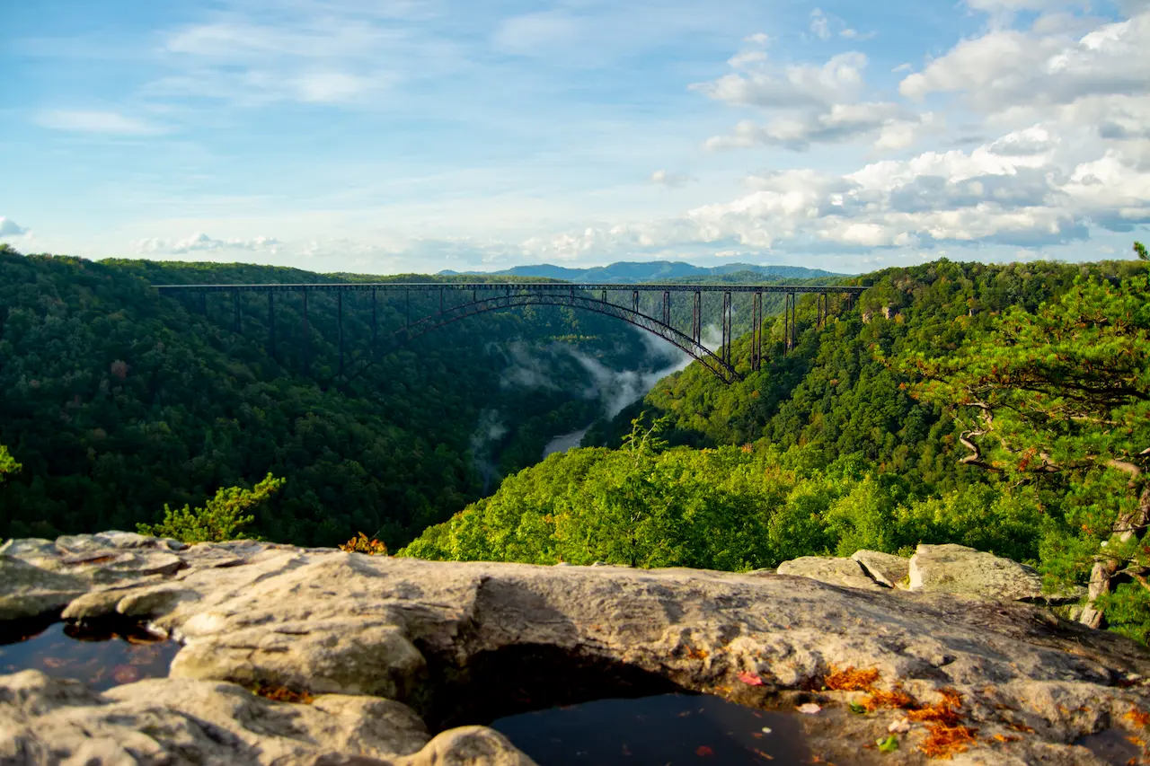 Panoramic view of the New River Gorge Bridge stretching across a wide, green valley, seen from a rocky overlook with water pools in the foreground.