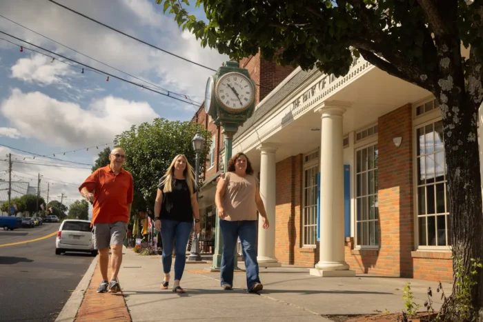 Three people walk together on a sunny day along a small-town sidewalk, passing by a brick building with white columns and a vintage-style clock.
