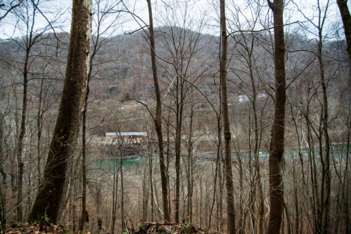 A small yellow building, a rusting bridge, and white dwellings a hill next to a river, peeking out through barren trees.