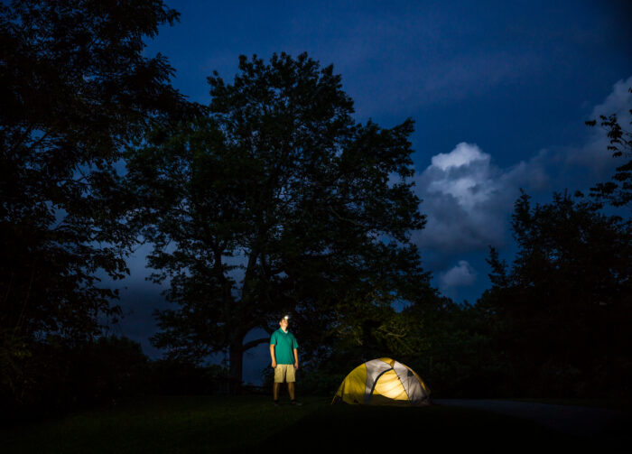 man standing outside of a lit tent with a headlamp glowing in a forest at night