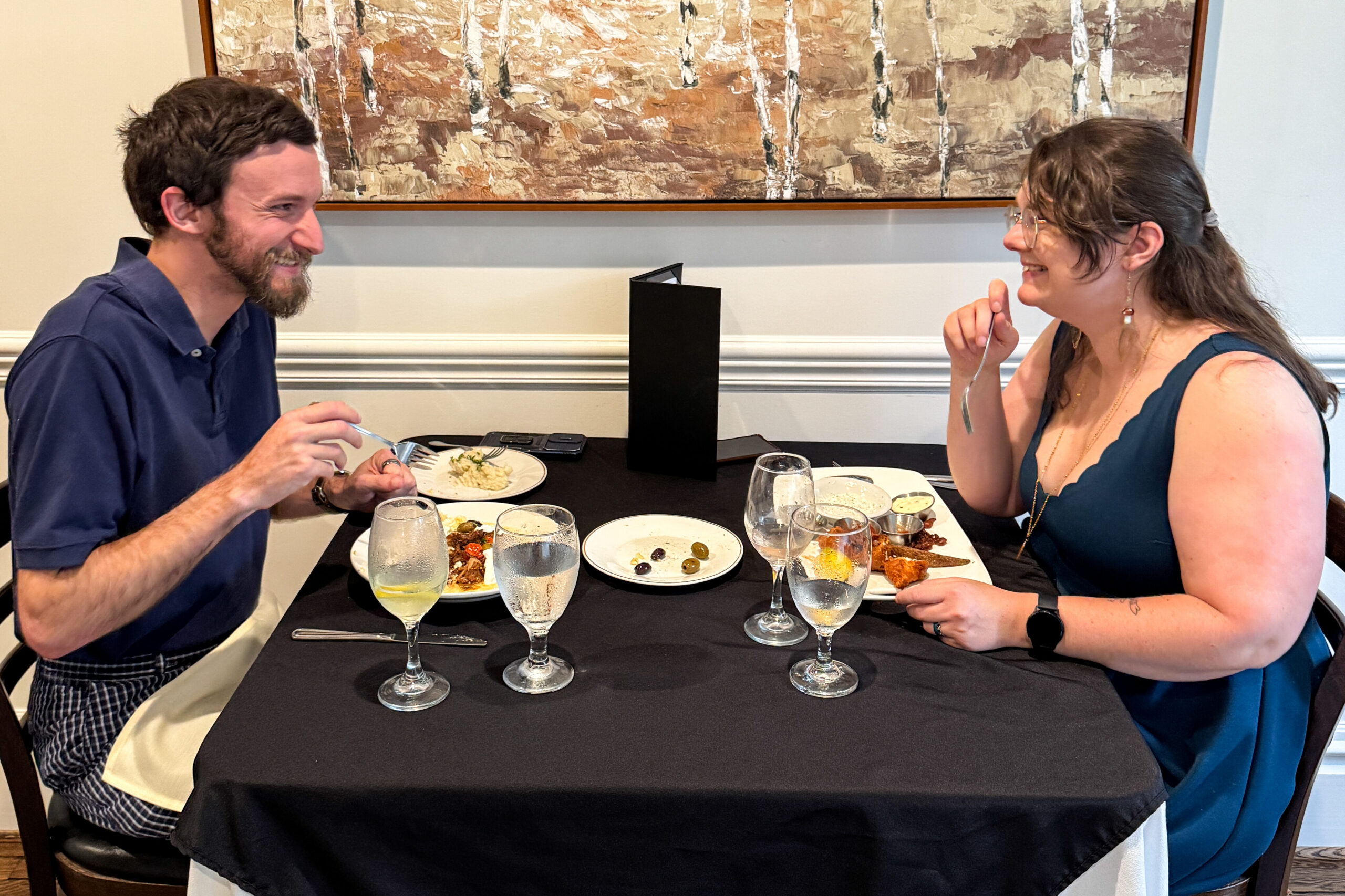 A man and woman holding forks, laughing and smiling with plates of food while sitting at a dining table