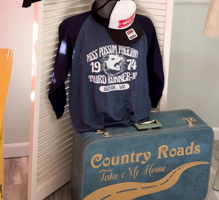 Vintage navy sweatshirt with "Miss Possum Pageant 1974 Third Runner-Up" text, a trucker hat, and a blue "Country Roads" suitcase.