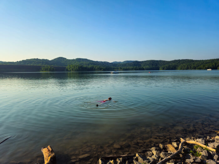 Calm lake with swimmer in red among gentle ripples, surrounded by distant tree-covered hills under clear blue sky.