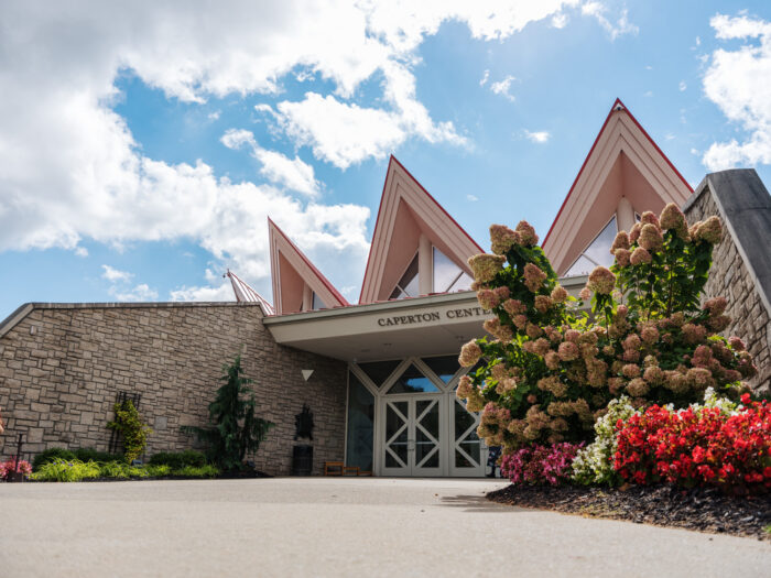 Caperton Center building with distinctive angular pink roofline, stone walls, and colorful flower gardens under a partly cloudy sky.