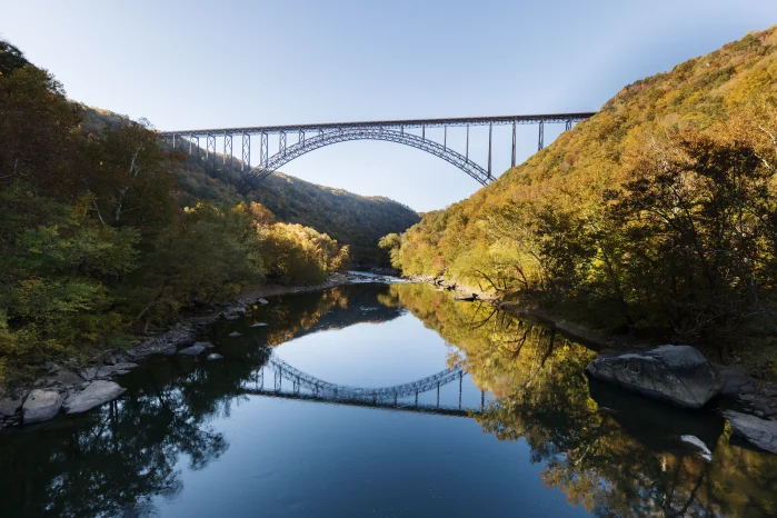 A steel arch bridge spans a gorge over a calm river, reflecting in the water, surrounded by autumn foliage.