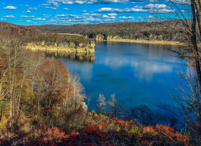 Aerial view of a blue lake surrounded by autumn foliage, limestone cliffs, and forested hills under a partly cloudy sky.