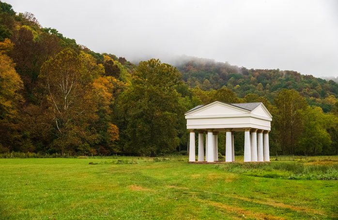 A white Greek-revival pavilion with columns stands alone in a green meadow, backed by misty autumn forest hills.
